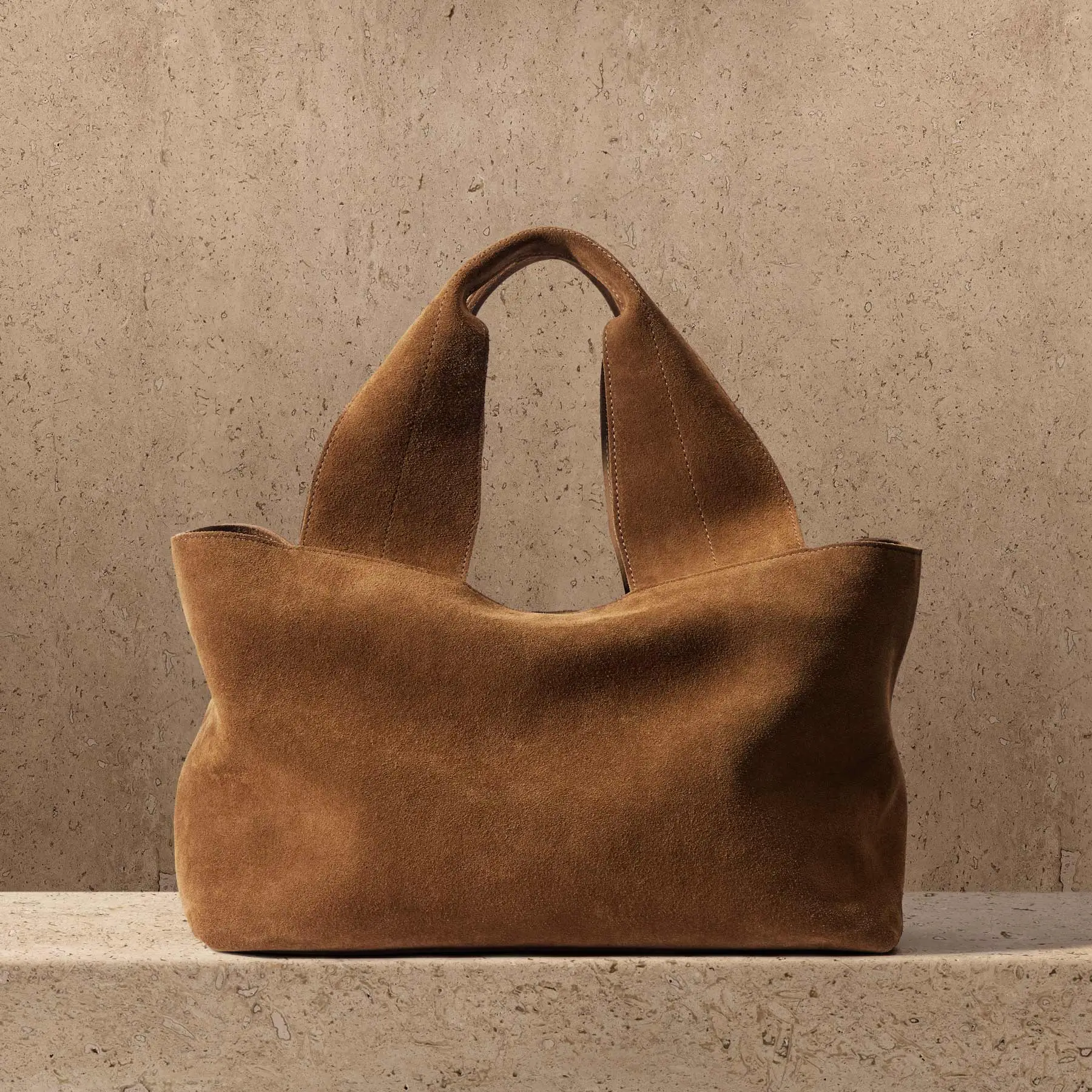 Soft-structured James Perse suede tote bag in warm brown tone, featuring wide integrated handles and a minimalist silhouette, set against a textured stone backdrop