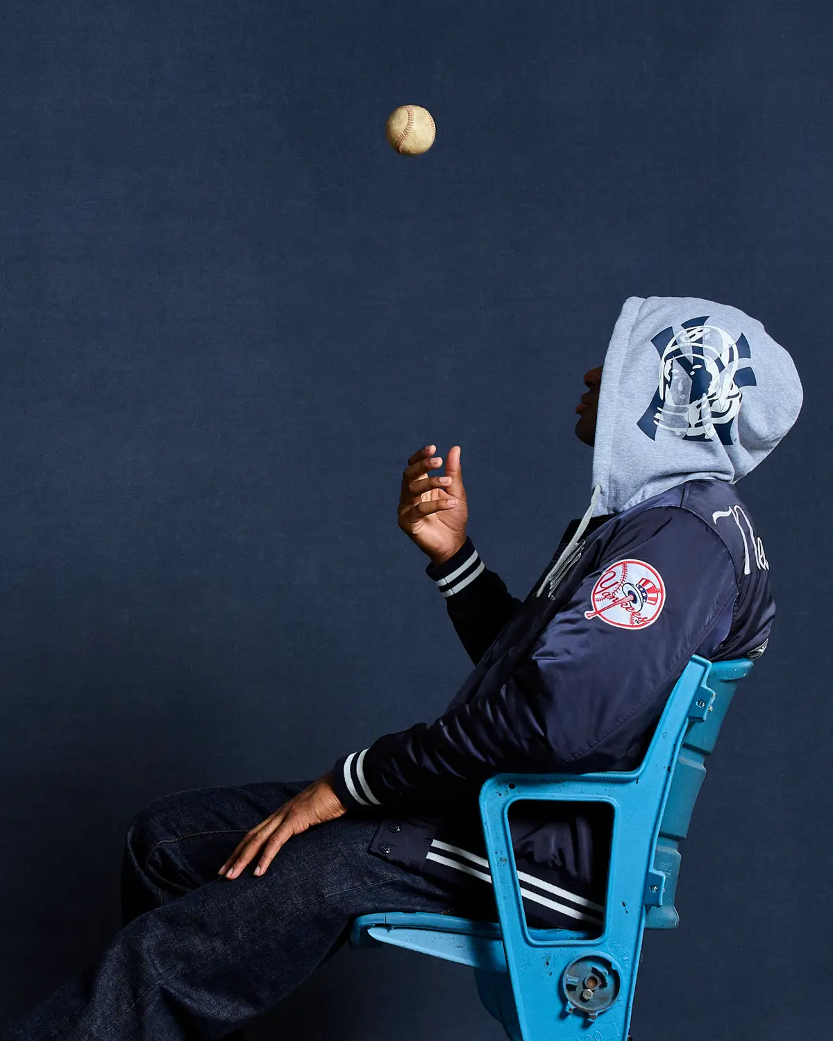Side profile of a model seated on a blue stadium chair wearing a navy New York Yankees varsity-style jacket layered over a grey hooded sweatshirt with graphic detailing, casually tossing a baseball in the air against a deep blue backdrop