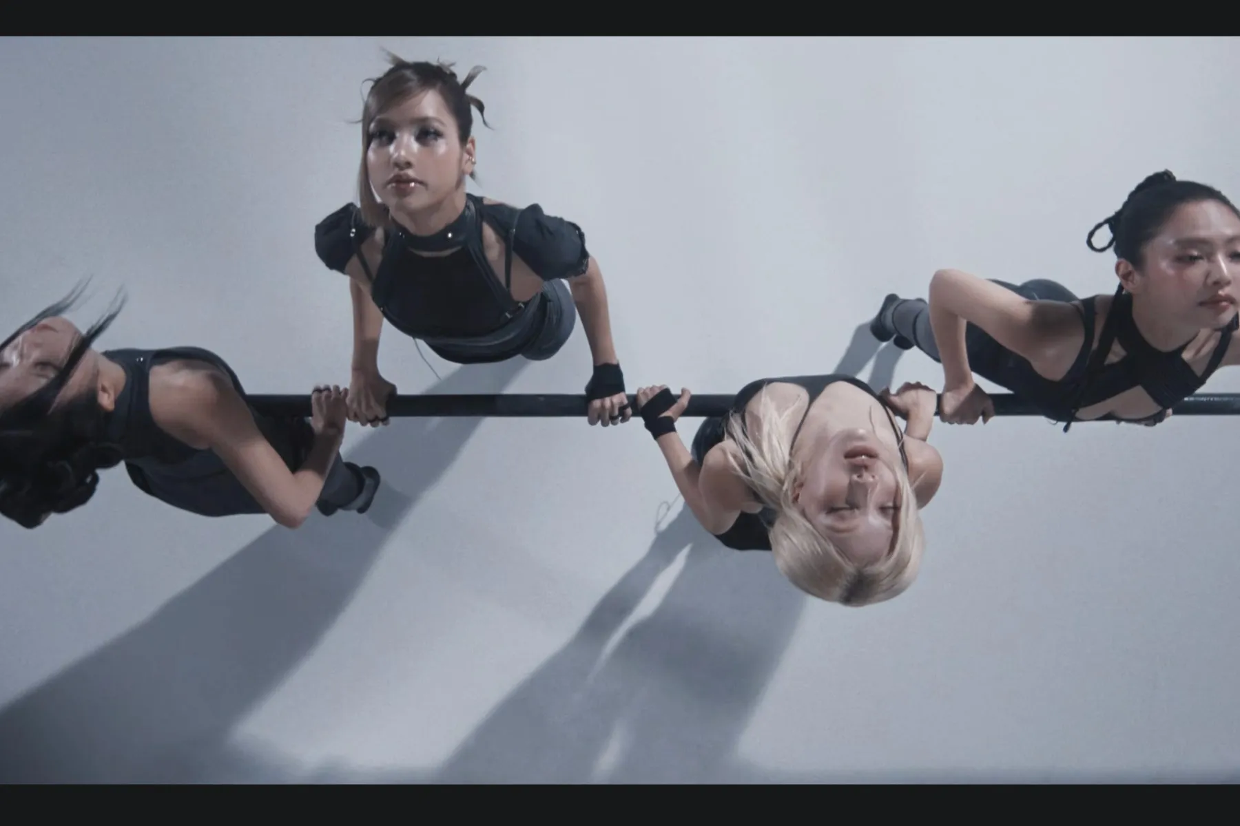 Overhead shot of BLACKPINK members dressed in coordinated black performance outfits, gripping a horizontal bar against a stark white backdrop, forming a symmetrical, high-intensity choreography pose for the “GO” era teaser