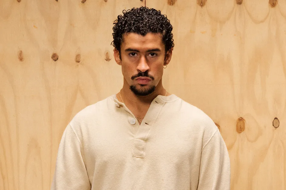 Portrait of Bad Bunny standing against a light wooden backdrop, wearing a beige henley shirt with a serious expression, highlighting the Puerto Rican artist whose adidas BadBo design inspired Team Puerto Rico’s World Baseball Classic cleats