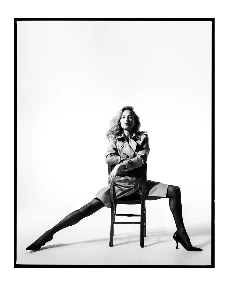 Black-and-white portrait of Kate Moss seated on a wooden chair wearing a classic Burberry trench coat, part of the “The Trench: Portraits of an Icon” campaign photographed by Tim Walker