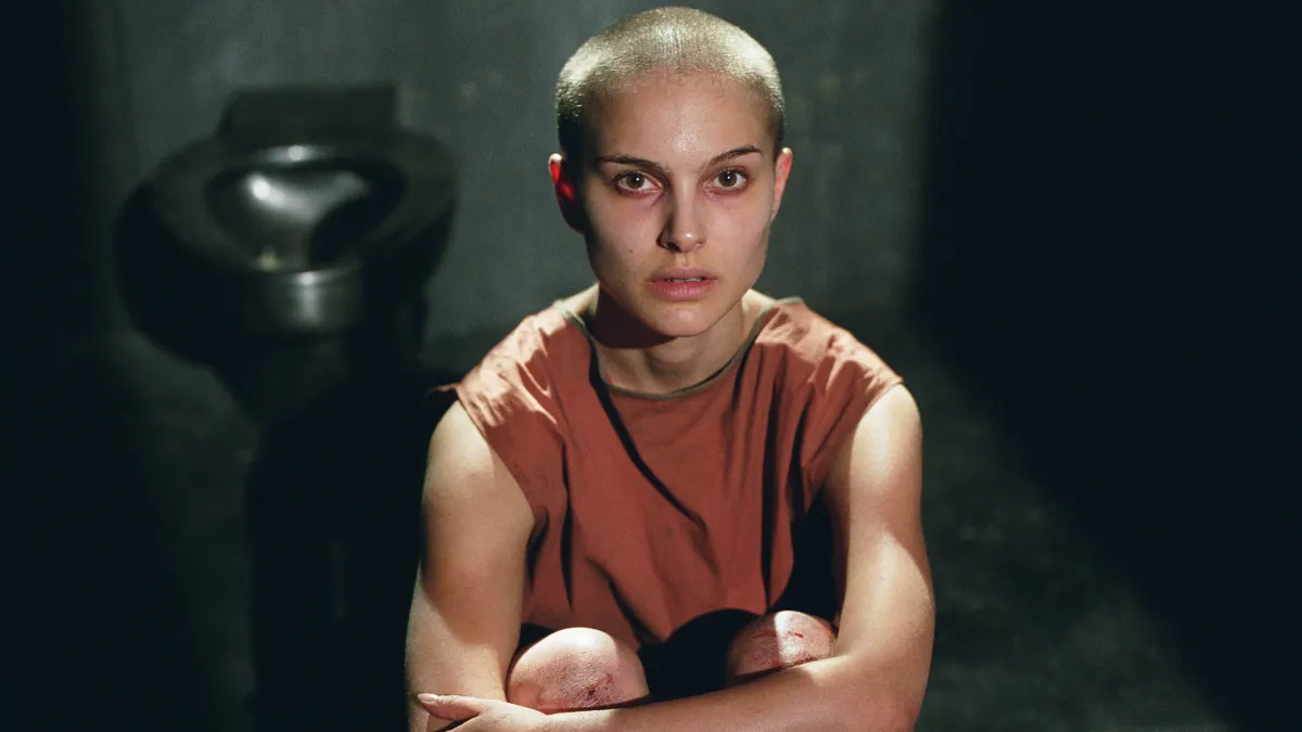 Natalie Portman as Evey Hammond in V for Vendetta, seated with a shaved head in a dimly lit prison cell, conveying vulnerability and transformation during the film’s pivotal interrogation sequence
