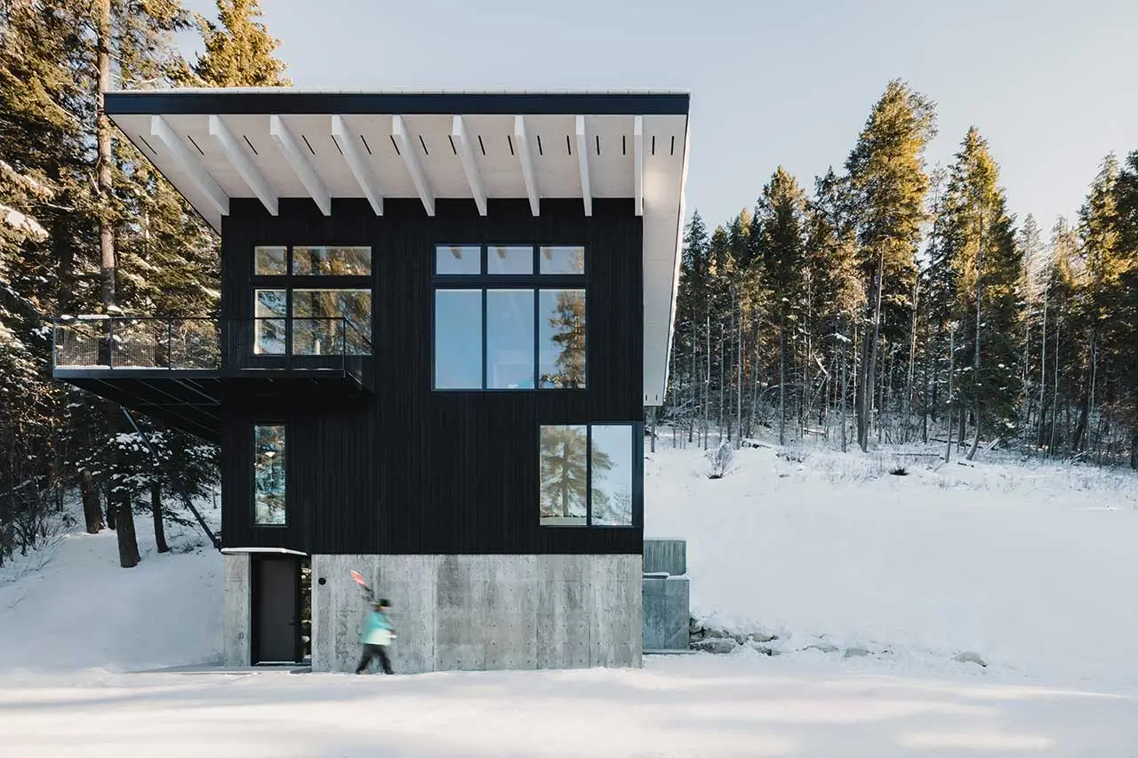 Modern black timber cabin with large windows and white overhanging roof perched on a concrete base in snowy forest near Golden, British Columbia, with a person walking outside the Columbia River Valley Lookout by Twobytwo Architecture Studio