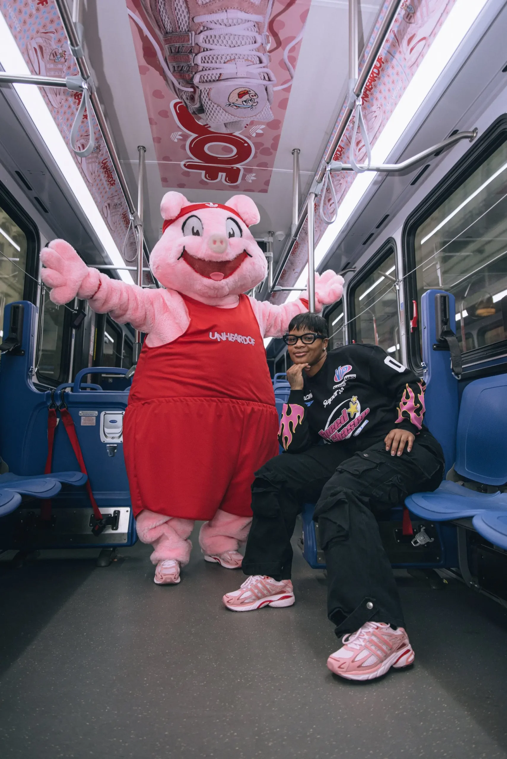 Person wearing pink Unheardof Piggy Runner sneakers sits inside a subway car beside a large pig mascot in a red outfit, with branded campaign graphics overhead, highlighting the playful storytelling of the Piggy Runner 2.0 collection