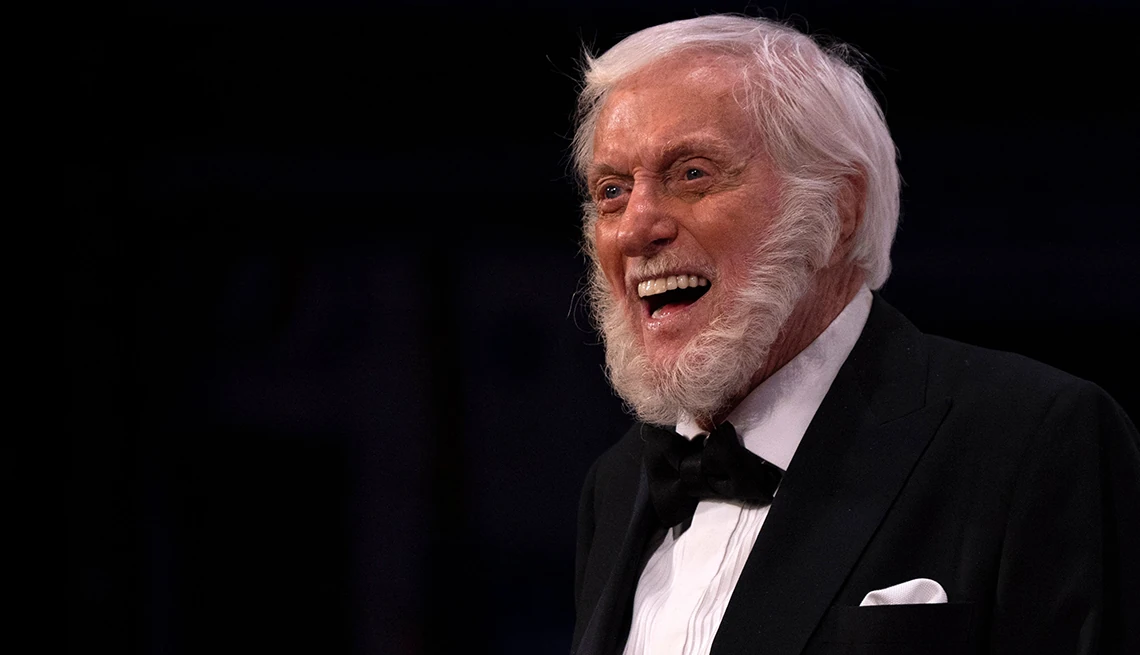 Dick Van Dyke in a tuxedo with a full white beard, smiling broadly on stage under soft lighting, captured mid-expression with an energetic and joyful presence