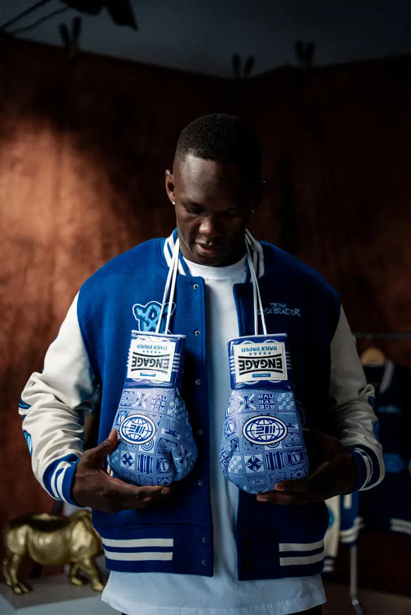 Man wearing a blue varsity jacket holds a pair of blue Engage boxing gloves featuring intricate Adinkra-inspired patterns, highlighting the Daily Paper collaboration’s blend of heritage symbolism and combat sport design
