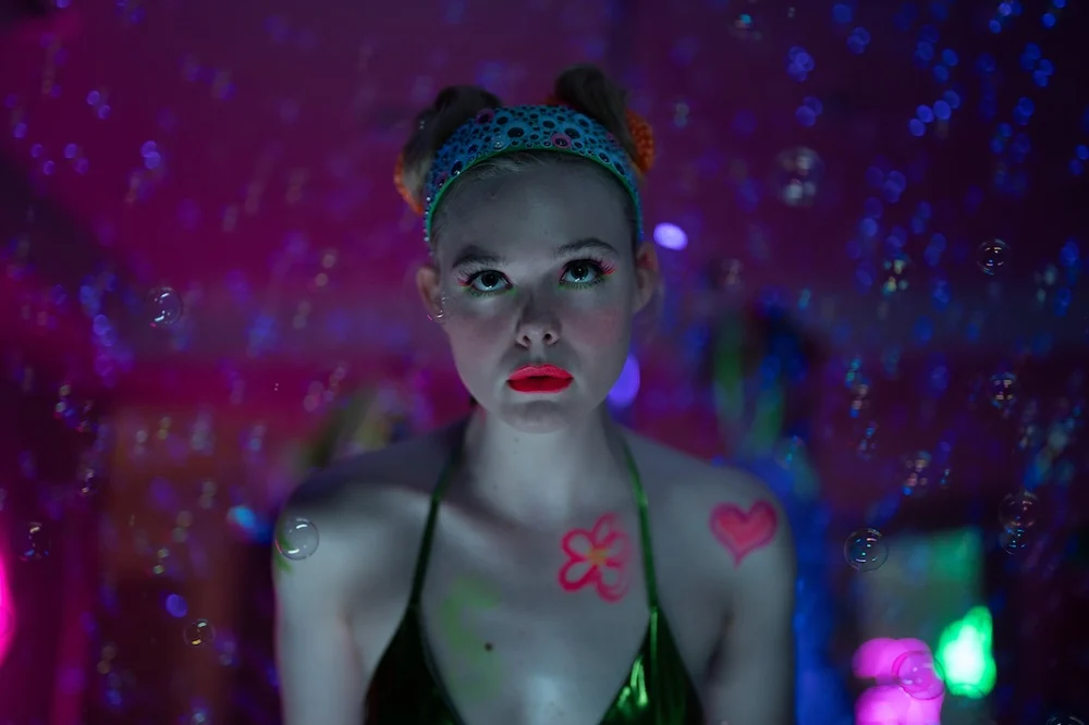 A softly lit, neon-saturated portrait of a young woman gazing upward, her expression suspended between curiosity and unease. She wears a glittering headband with her hair styled into two small buns, while fluorescent body paint—hearts and playful motifs—marks her shoulders and chest. The environment glows with iridescent pinks, purples, and blues, punctuated by floating bubbles that drift across the frame, creating a dreamlike, almost artificial atmosphere that evokes digital fantasy and performative identity