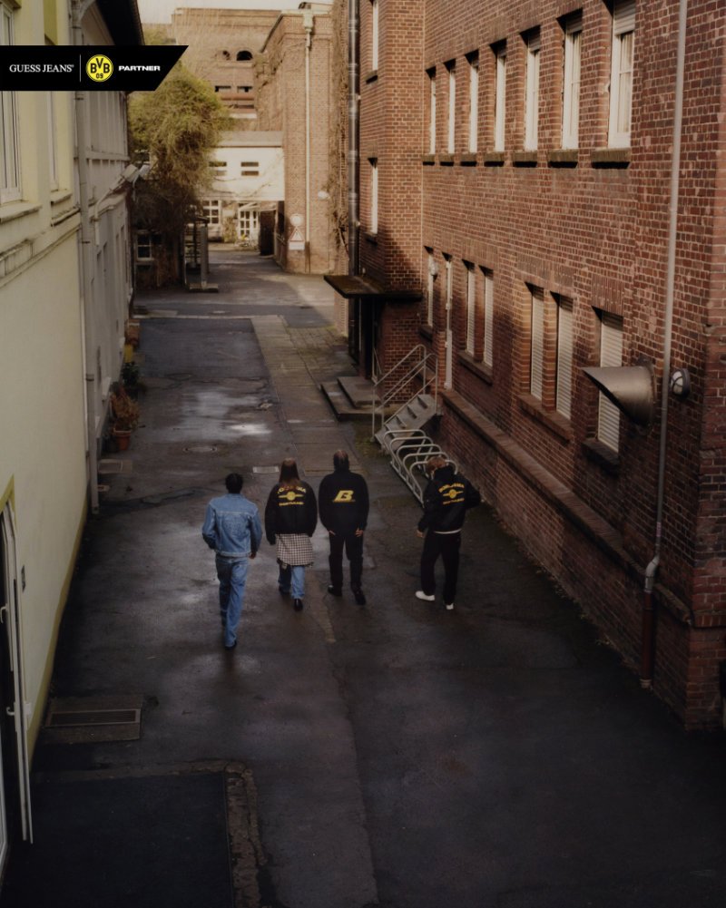 Four supporters wearing GUESS JEANS x Borussia Dortmund jackets and denim walk down a narrow city alley in Dortmund, highlighting the collaboration’s streetwear aesthetic inspired by BVB fan culture