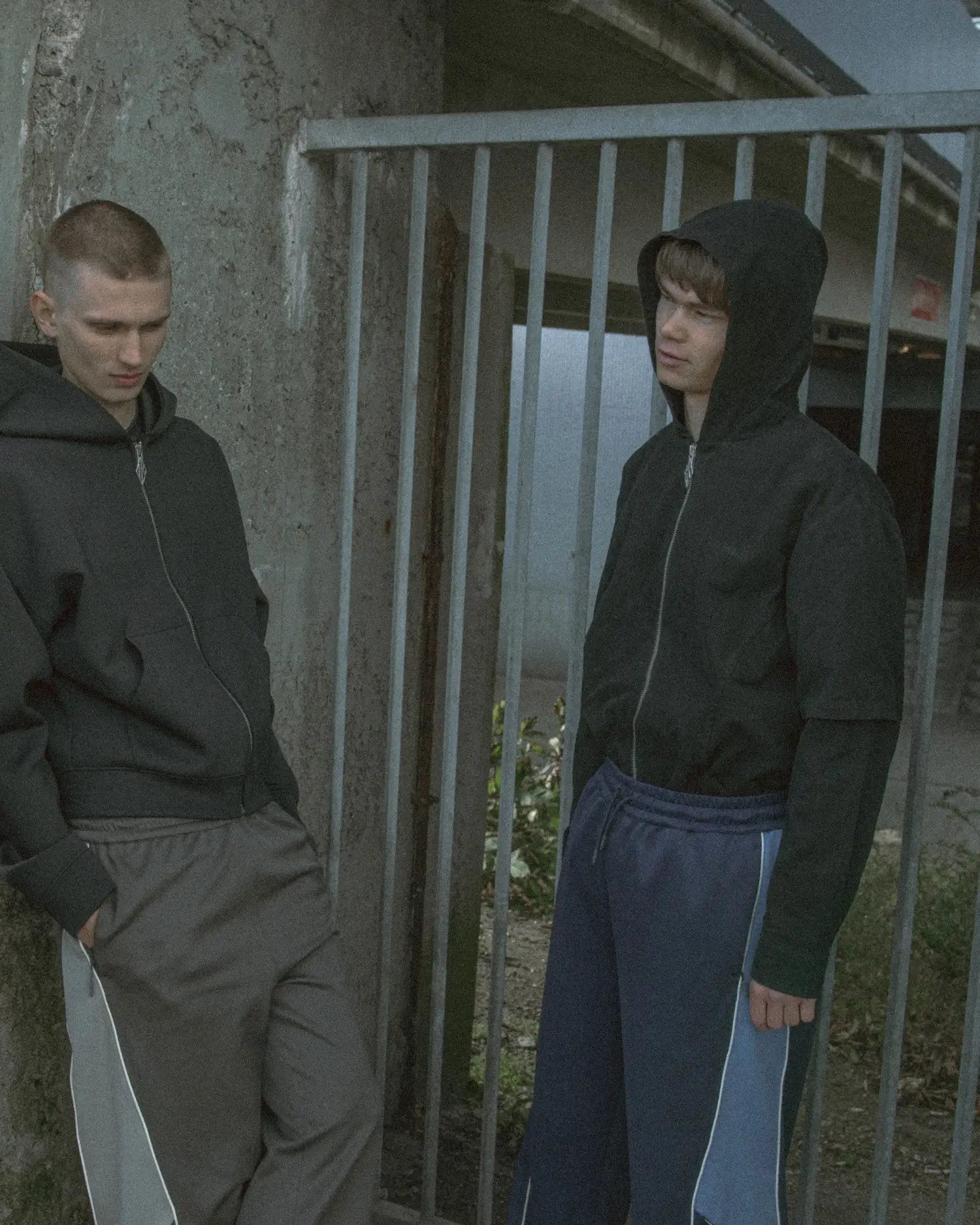 Two models stand in an urban outdoor setting beside a concrete wall and metal railing, wearing Han Kjøbenhavn Pre-AW26 hooded zip jackets and relaxed track pants with subtle stripe detailing, conveying a muted, football-inspired street aesthetic