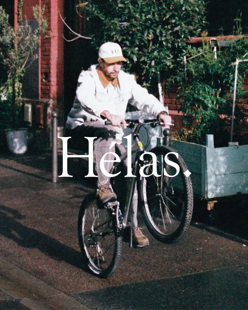 Skater riding a bicycle through a sunlit residential street wearing a light Hélas outfit and cap from the Hélas Spring 2026 collection, with the Hélas logo overlayed across the image