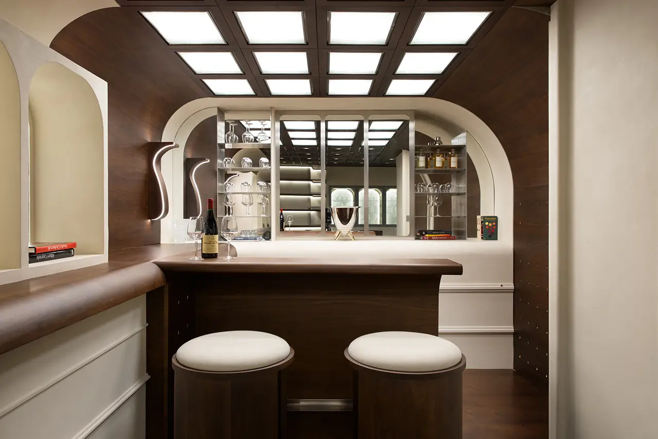 Custom-designed minimalist interior featuring a warm wood-paneled ceiling with integrated square lighting, a central handcrafted bar counter with rounded edges, and mirrored shelving units displaying glassware and spirits, creating a refined private office or lounge environment defined by symmetry, soft illumination, and architectural detailing