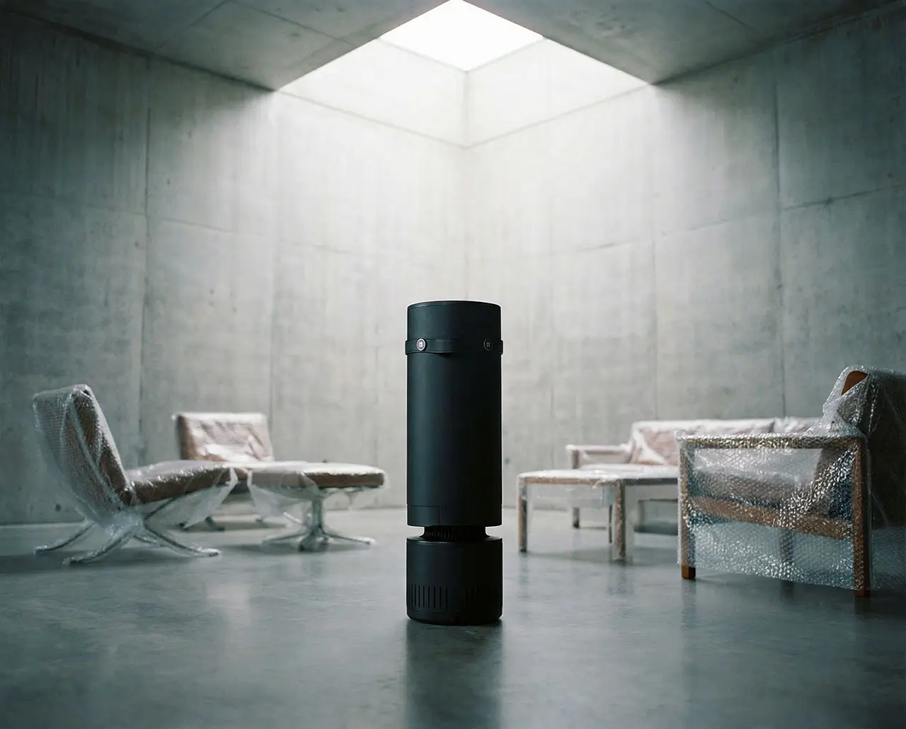 Black cylindrical smart space heater standing in the center of a minimalist concrete room under a skylight, surrounded by modern lounge chairs wrapped in protective plastic