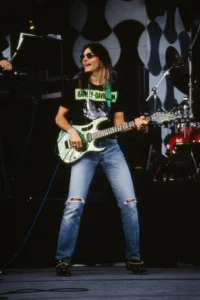 Steve Vai performing live on stage with a green electric guitar, wearing sunglasses, a Harley-Davidson T-shirt, and ripped jeans during a rock concert performance