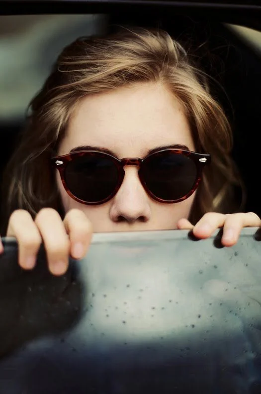Close-up of a woman wearing round tortoiseshell sunglasses in a vintage-inspired minimalist design, gripping a car window frame, with soft natural lighting emphasizing the understated haute aesthetic of premium eyewear