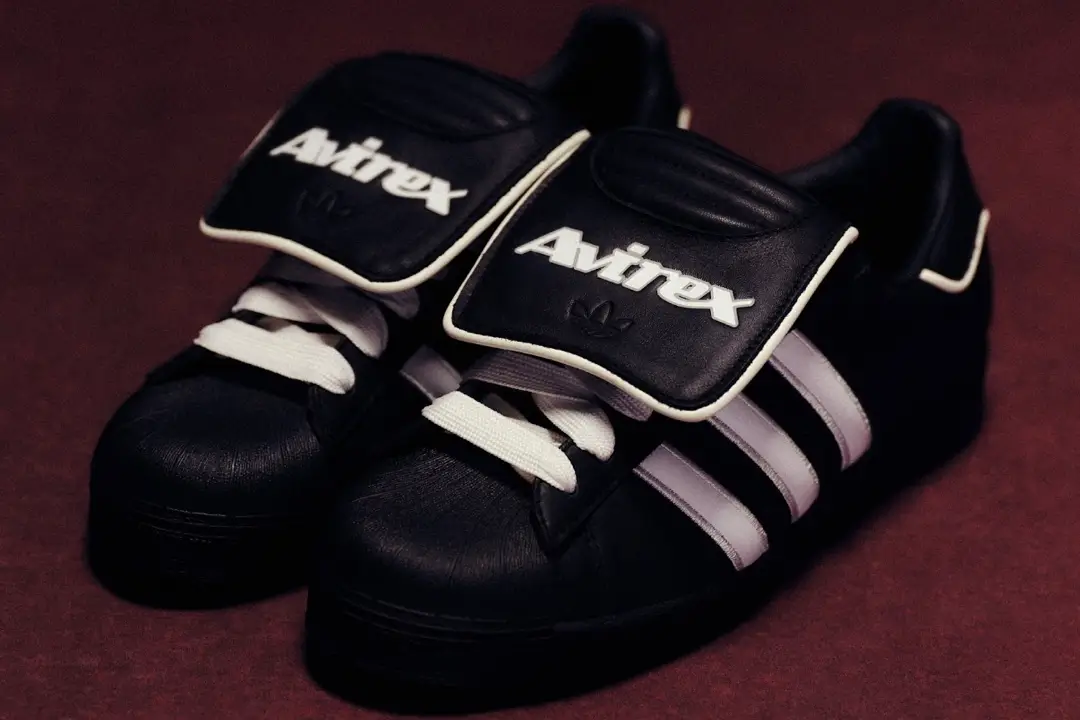 AVIREX x adidas Superstar 82 “Core Black” sneakers featuring premium black leather uppers, white three-stripe branding, shell toe design, and oversized AVIREX-branded tongue flaps with white laces