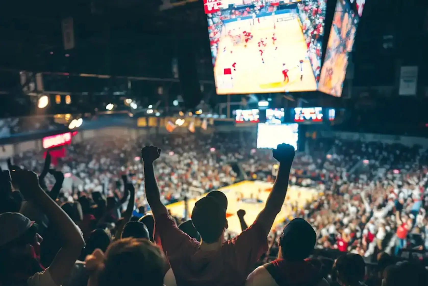 Crowd of basketball fans inside a packed arena cheering with raised arms as a high-energy game unfolds on the court below, illuminated by bright scoreboard screens overhead