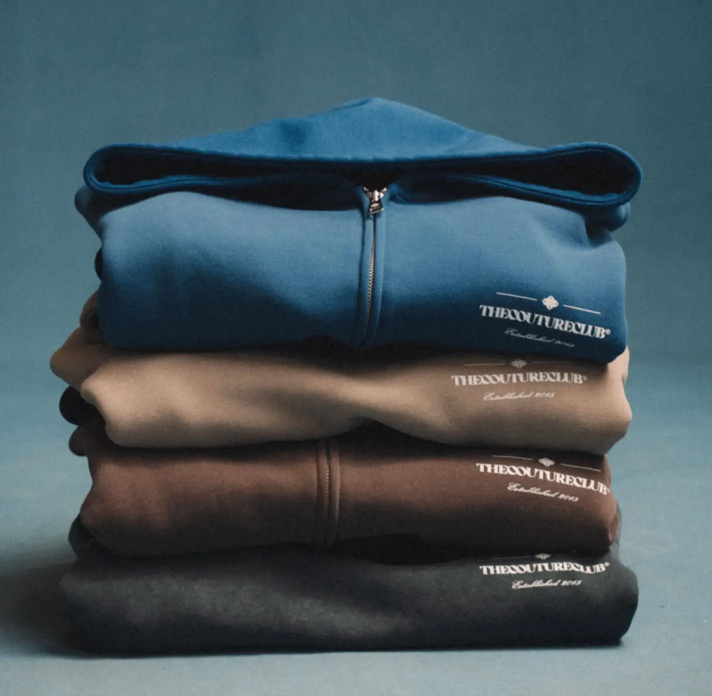 Folded stack of The Couture Club zip-up hoodies in blue, beige, brown, and black, neatly layered against a minimalist backdrop, showcasing soft-touch fabric and embroidered logo branding
