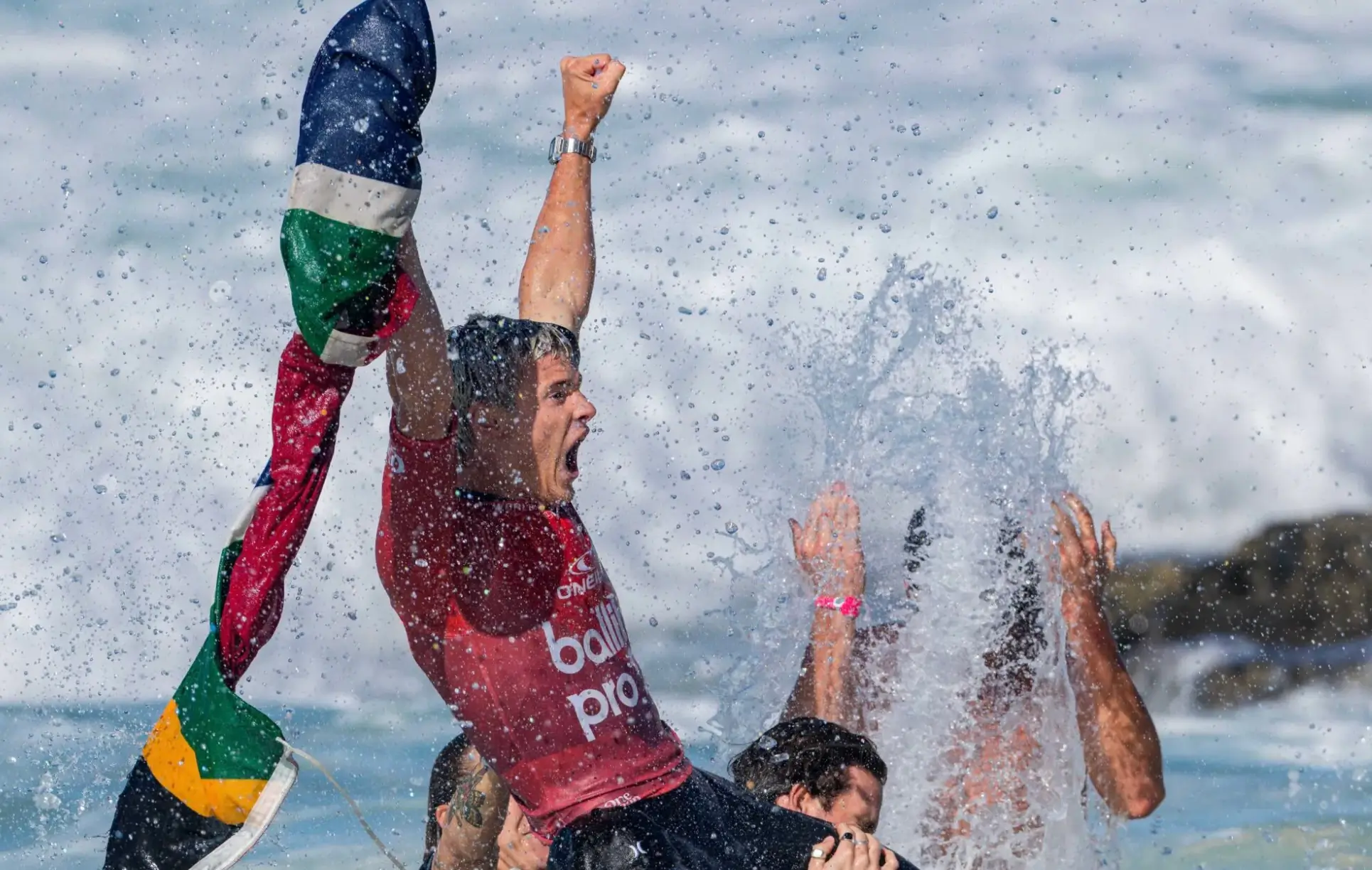Professional surfer in a red competition jersey celebrates victory in the ocean, raising his fist while holding a national flag as water splashes around him and fellow competitors cheer nearby