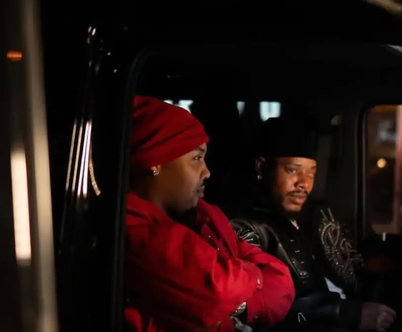 left G hero as right side Fetty Wap inside a dimly lit vehicle at night, one in a red headwrap and matching jacket, the other in a black cap and leather jacket, both facing forward in a reflective, subdued moment that mirrors the introspective tone of the song