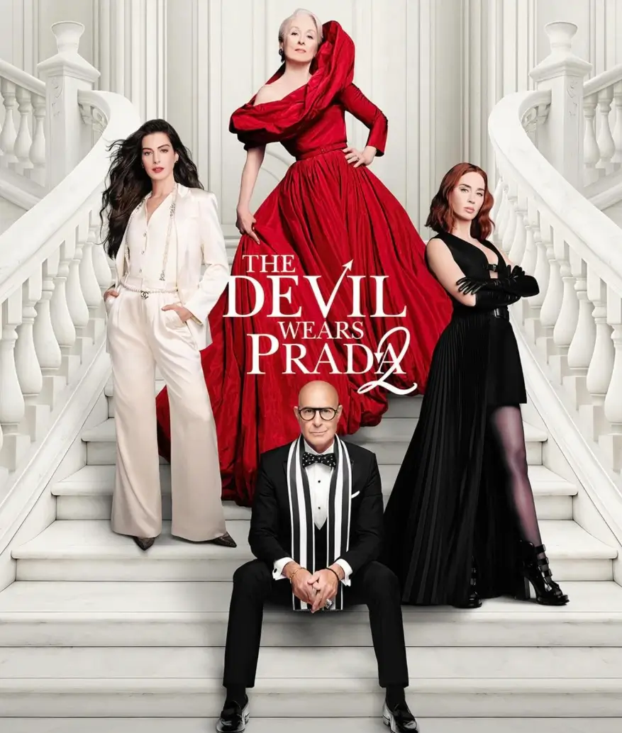 Promotional poster for The Devil Wears Prada 2 featuring a central figure in a dramatic red couture gown standing atop a grand white staircase, flanked by two sharply dressed women in contrasting tailored looks—one in an ivory suit and the other in a black pleated ensemble—while a suited man sits below, creating a symmetrical composition that emphasizes power, hierarchy, and high-fashion authority