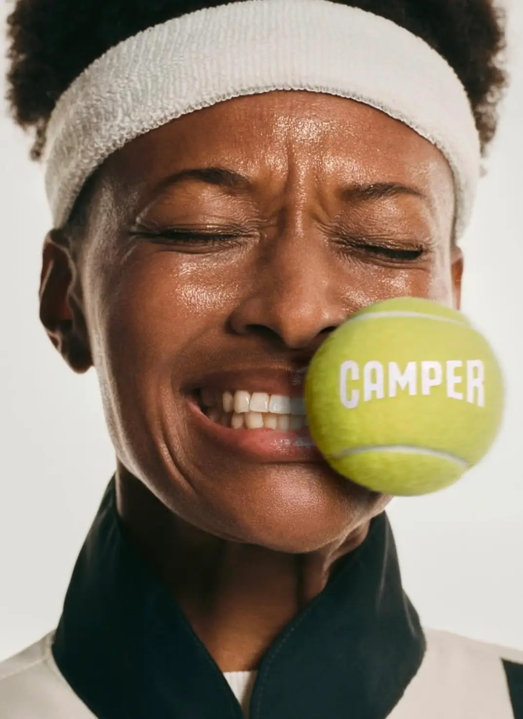 Close-up portrait of a model wearing a white tennis headband, eyes squeezed shut as a bright yellow tennis ball stamped with Camper branding presses against their face, capturing the playful, offbeat humor of the “Crazy for Tennis” campaign
