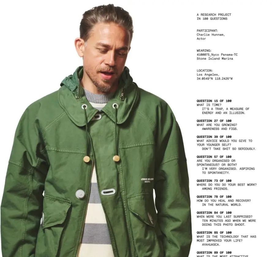 Charlie Hunnam wearing a green Stone Island Marina Nyco Panama-TC jacket with a foldaway hood, layered over a striped knit, photographed against a clean studio backdrop. To the right, printed text from “A Research Project in 100 Questions” lists his responses, including reflections on time, spontaneity, and personal growth, along with garment and location details