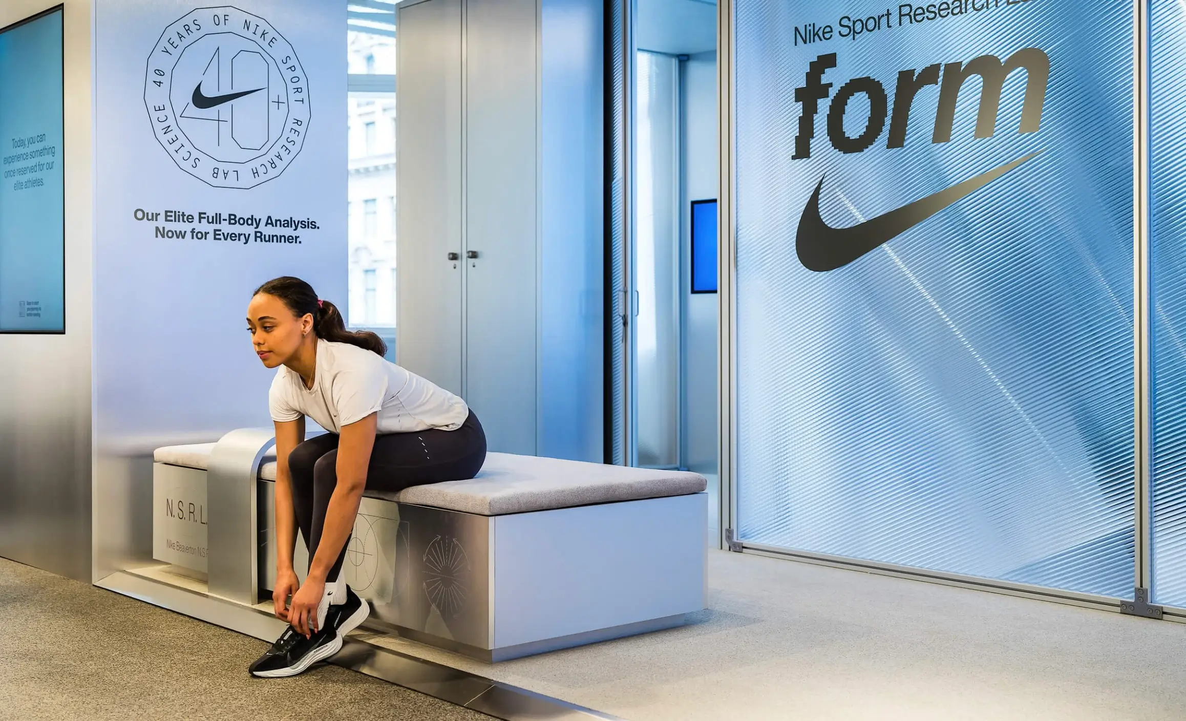 Visitor seated on an NSRL bench inside Nike Sports Research Lab pop-up, adjusting running shoes beneath signage reading “Our Elite Full-Body Analysis. Now for Every Runner,” with frosted glass panels displaying “form” branding in a clean, clinical retail environment