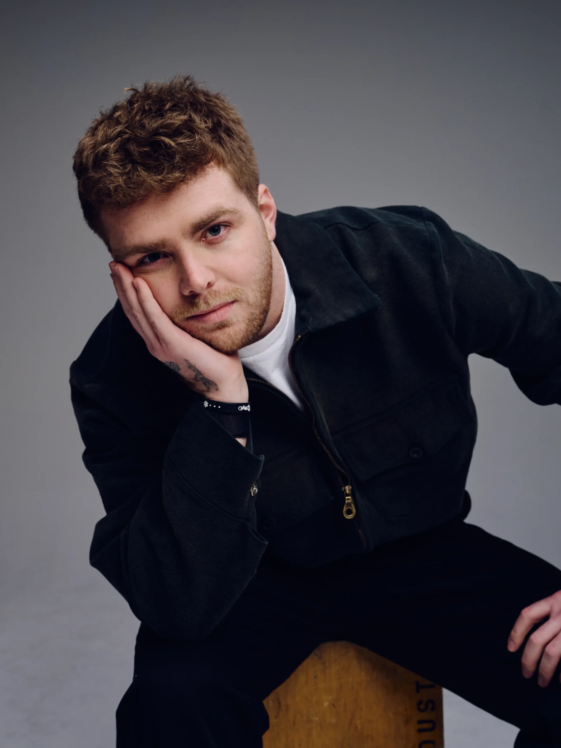 Portrait of Alex Warren leaning forward with his head resting on his hand, wearing a dark jacket over a white shirt against a neutral studio background
