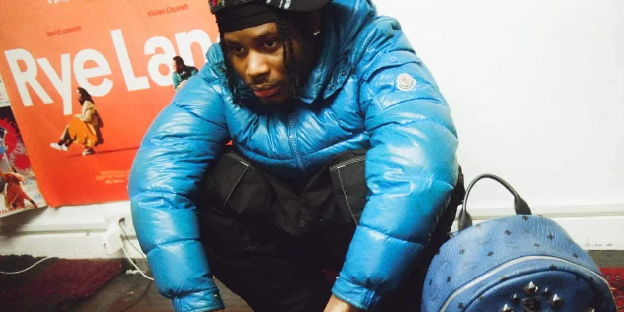 Jim Legxacy crouches in a blue puffer jacket and headphones, framed against a bold red wall poster, capturing the introspective and grounded mood surrounding “idk idk”