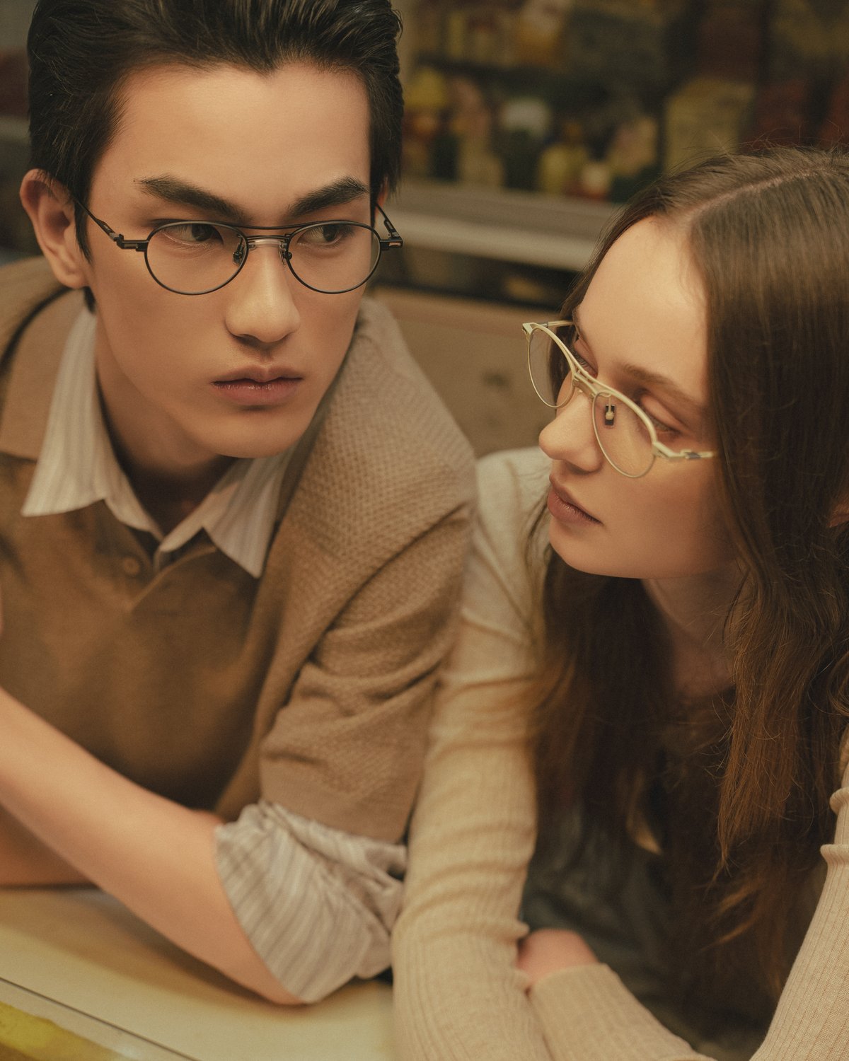 Two models wearing minimalist metal-frame eyewear, leaning closely in an intimate indoor setting, highlighting refined Japanese craftsmanship and understated design from the A. SOCIETY × Maison Kitsuné capsule