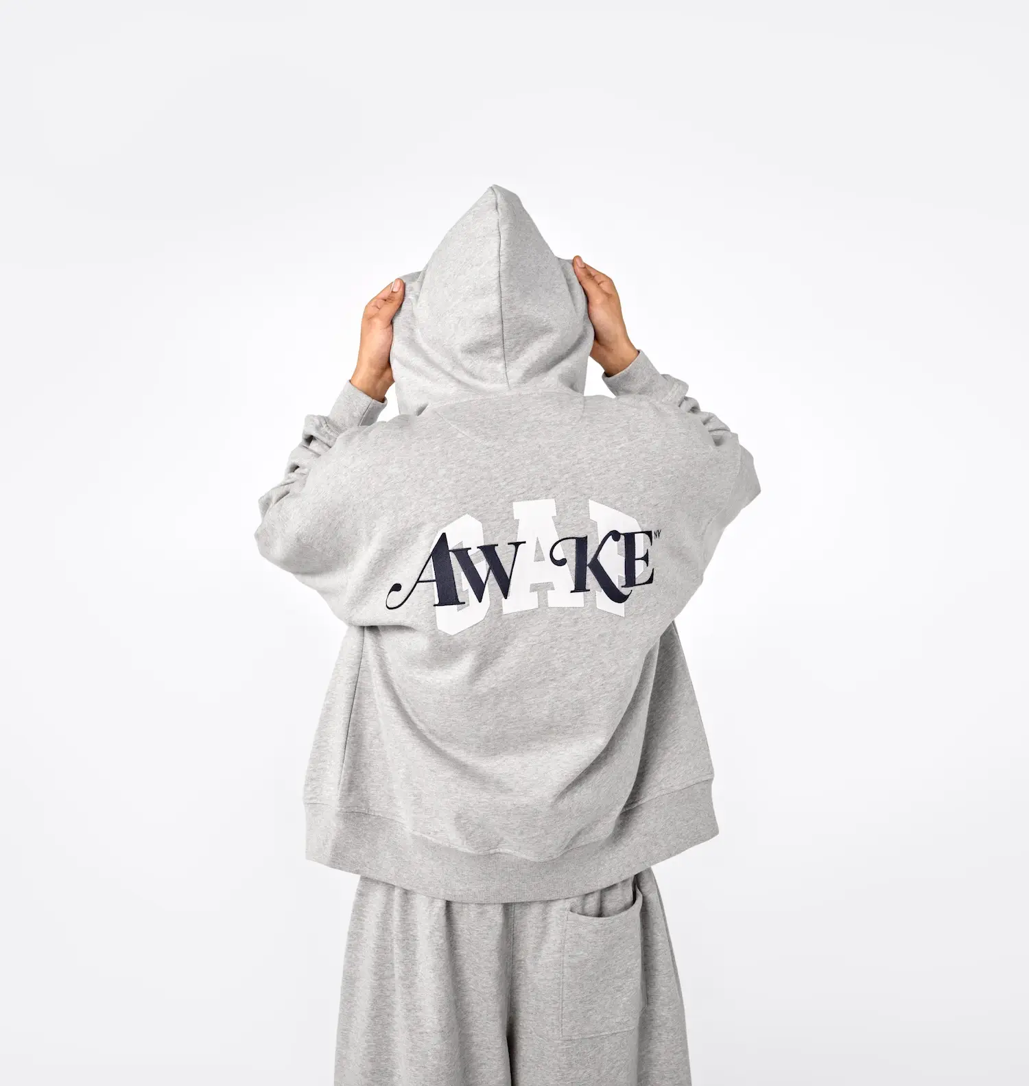 Back view of a model wearing a grey hoodie and matching sweatpants set, featuring layered “AWAKE” lettering over a large “GAP” logo across the back; the relaxed silhouette and minimal palette highlight co-branded graphic design and everyday streetwear styling from the Gap x Awake NY collection