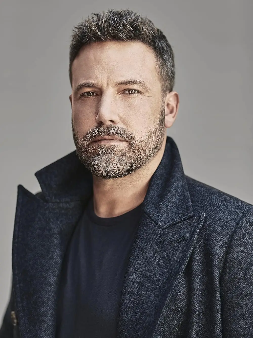 Portrait of Ben Affleck wearing a dark coat and black shirt against a neutral background, highlighting the Oscar-winning actor known for directing and starring in major Hollywood films