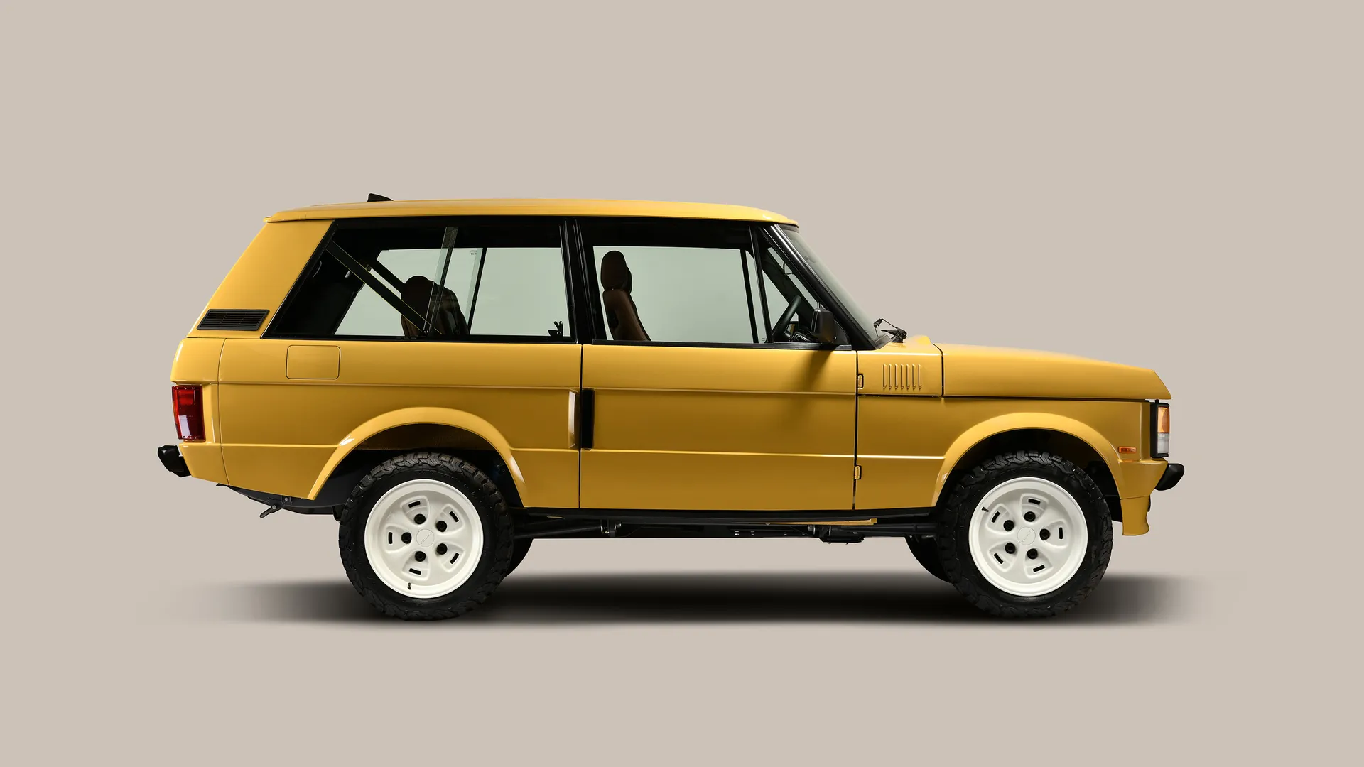 Side profile of a restored two-door Range Rover Classic in mustard yellow, featuring a clean, boxy silhouette, black trim, white steel wheels, and off-road tires against a neutral studio background