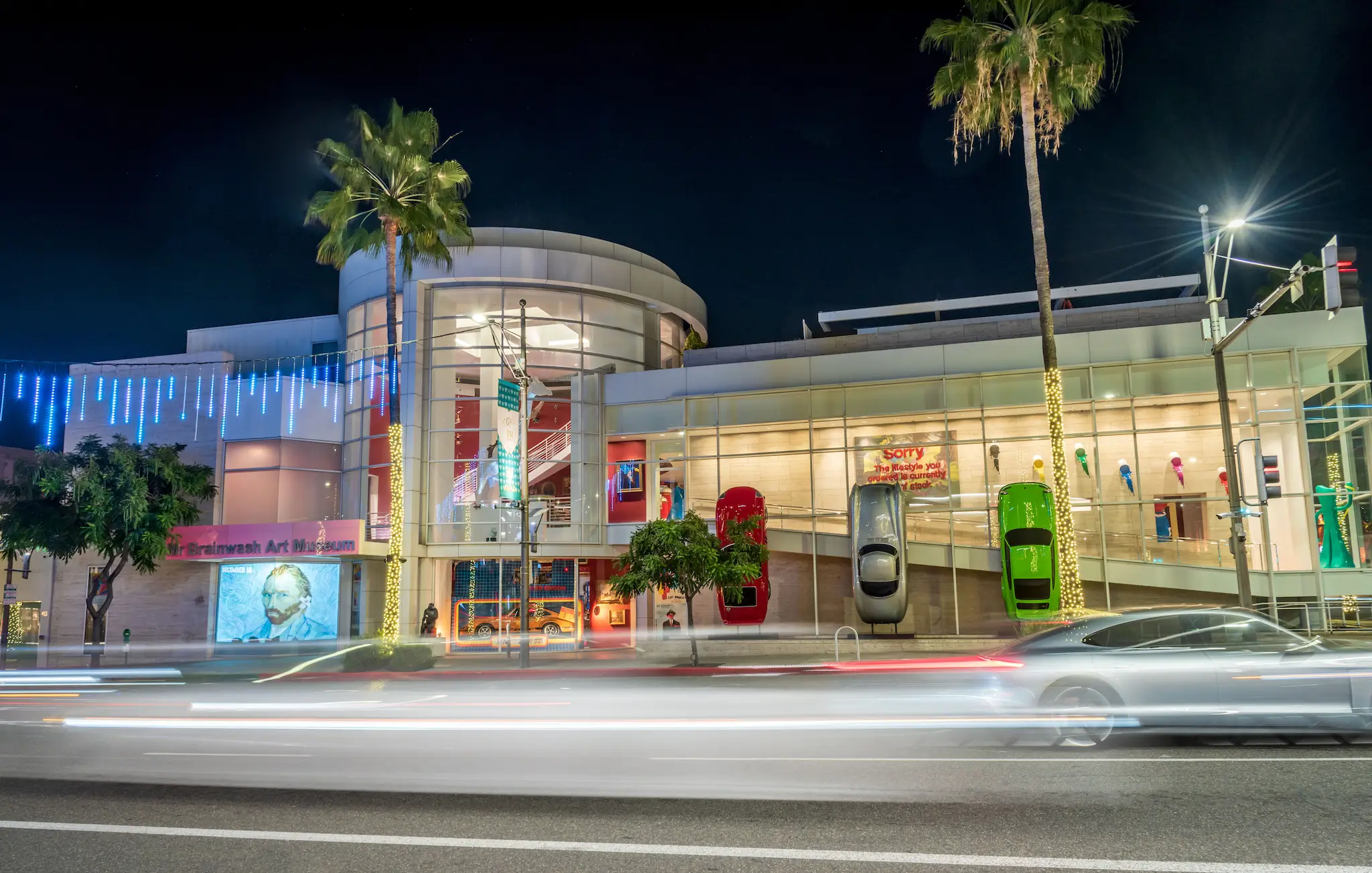 Night view of the Mr Brainwash Art Museum in Beverly Hills, Los Angeles, with illuminated palm trees, glass-front architecture, and colorful cars mounted vertically on the exterior wall