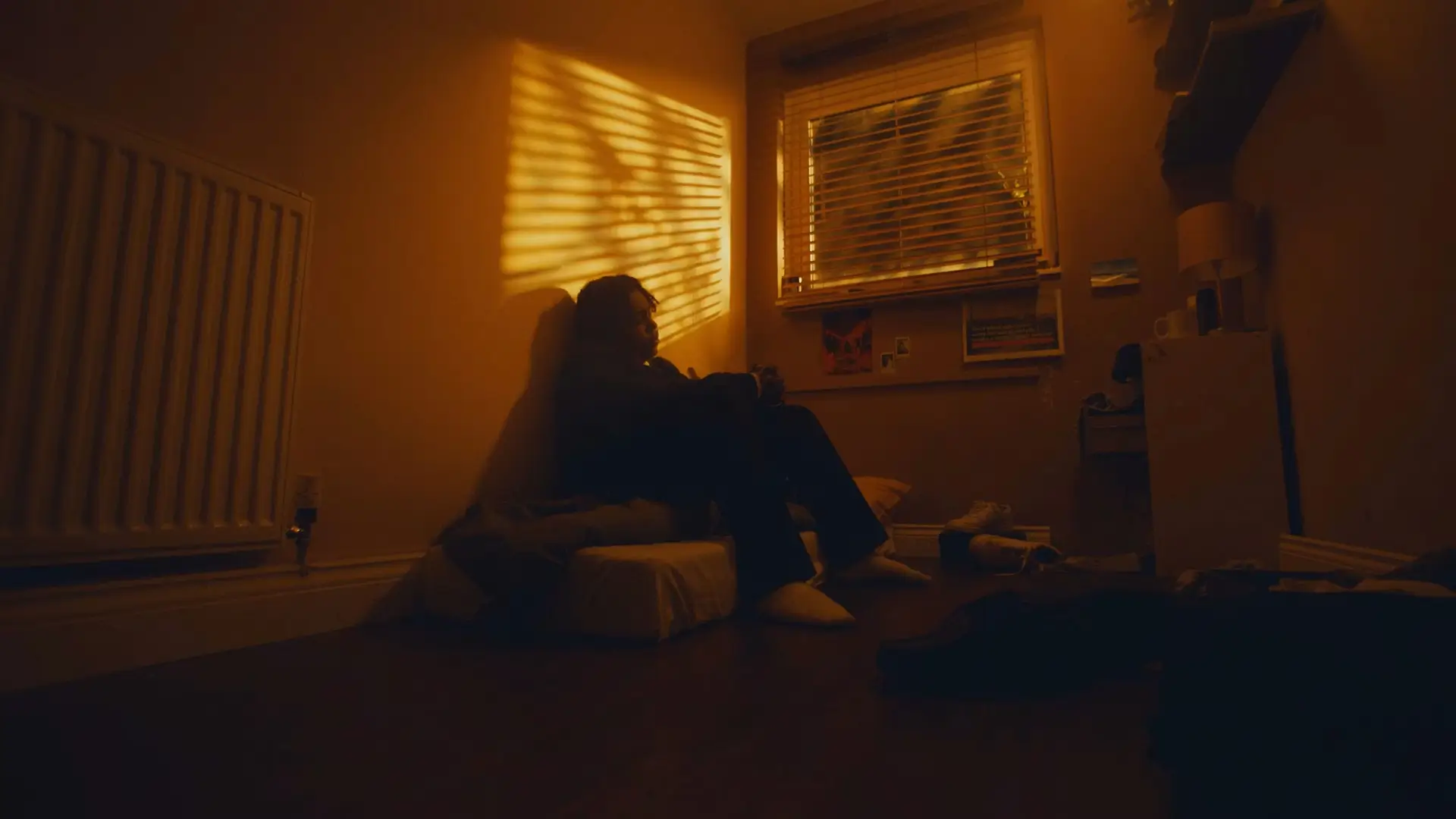 An adolescent sitting on a thin mattress on the floor in a dimly lit bedroom, warm light casting shadows through window blinds, illustrating the emotional impact of bed poverty and lack of proper sleeping conditions