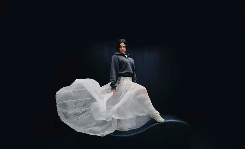 Rosalía posing against a dark studio background wearing a flowing white skirt and gray top while showcasing the New Balance 204L shoes during the brand’s campaign shoot