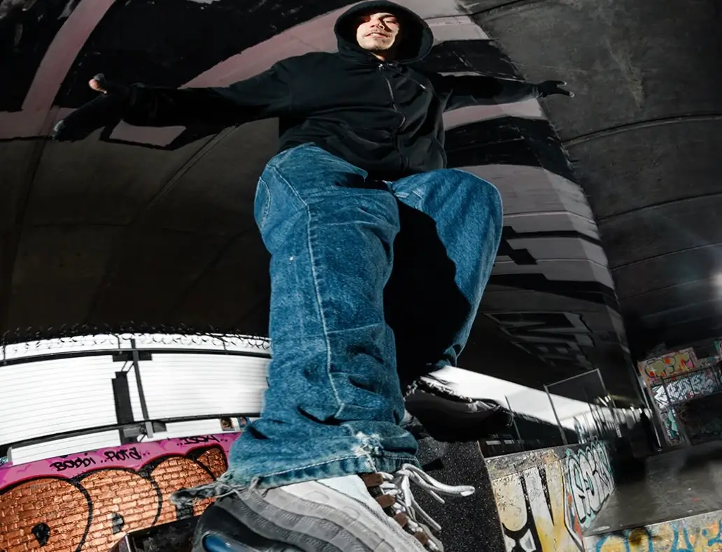 Low-angle shot of a person balancing on a ledge in an urban skate spot, wearing a black hoodie and loose blue jeans, with Nike Air Max 95 sneakers prominently visible in the foreground, set against a graffiti-covered concrete environment
