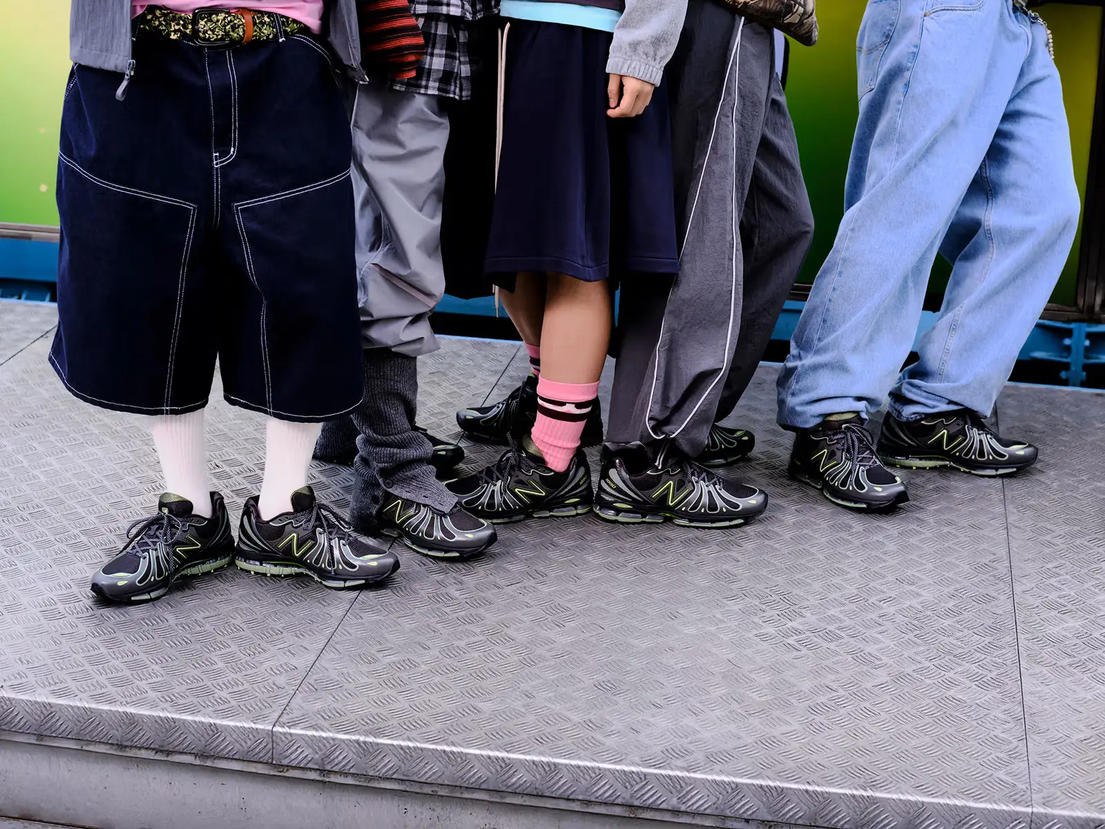 Group of people standing side by side wearing black and neon-accented New Balance 1890 sneakers, styled with loose denim, track pants, and streetwear socks during a lifestyle campaign shoot