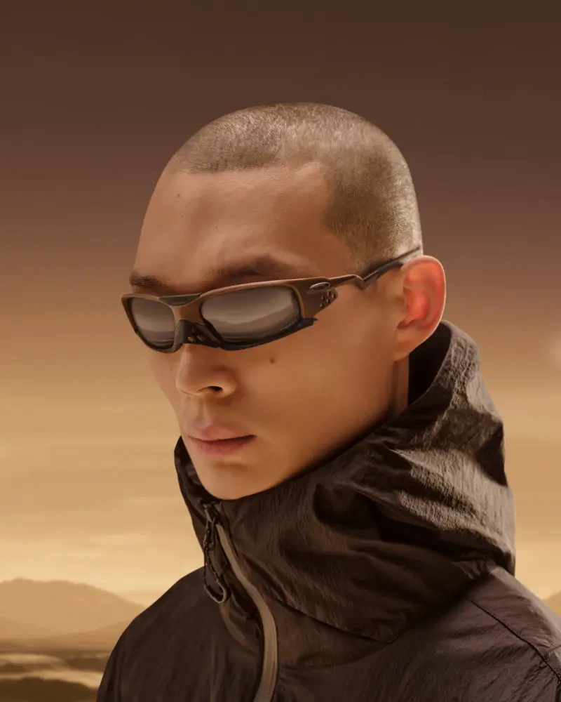 Model wearing Oakley Jacket eyewear with a matte brown wraparound frame and tinted lenses, paired with a technical high-collar field jacket, set against a muted desert landscape, highlighting the fusion of performance optics and minimalist Japan-inspired outdoor apparel design