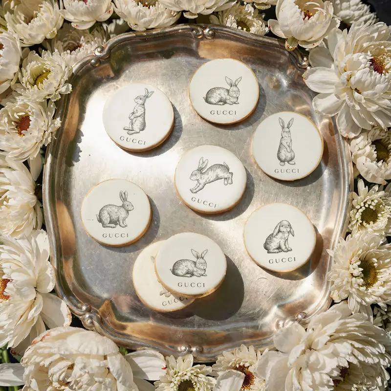 Gucci-branded round cookies with illustrated rabbits arranged on a silver tray, surrounded by white flowers in a refined spring haute presentation