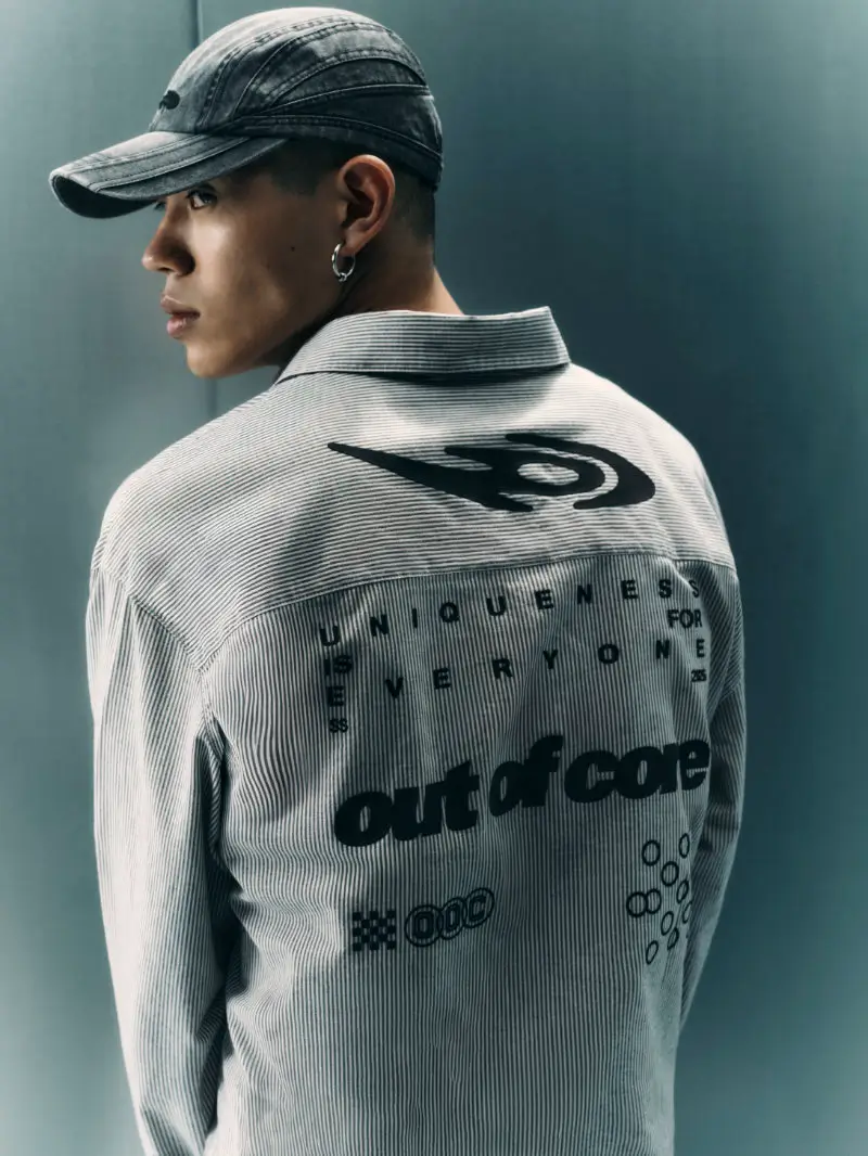 Model wearing a striped Bershka OUT OF CORE jacket with bold graphic text reading “UNIQUENESS FOR EVERYONE” and “out of core” across the back, styled with a fitted cap and earring, captured in a minimal studio setting with a muted teal backdro