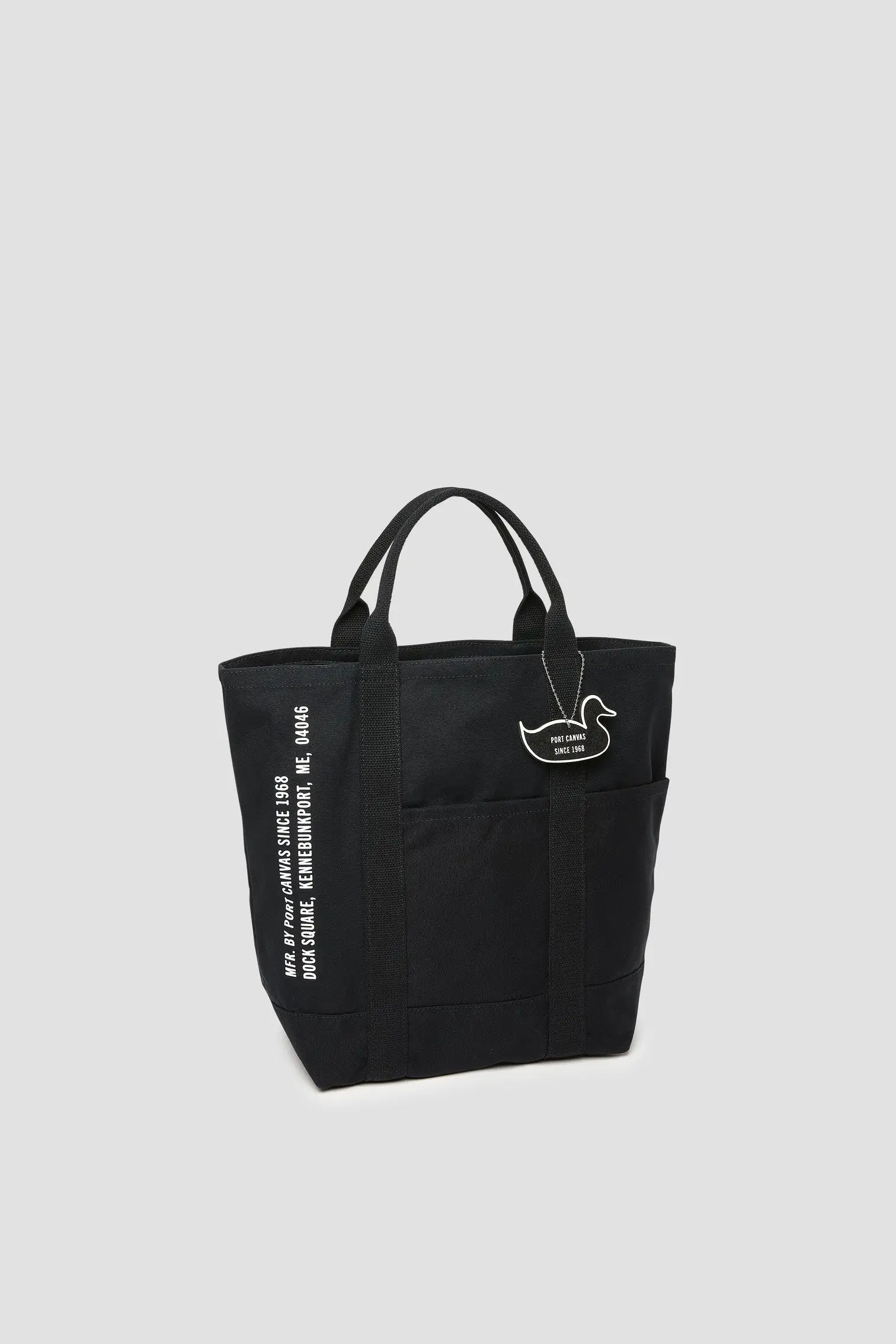 Black Port Canvas Double Utility Tote with dual exterior pockets, vertical logo print reading “Port Canvas Since 1968,” reinforced webbing handles, and duck-shaped branded tag on front