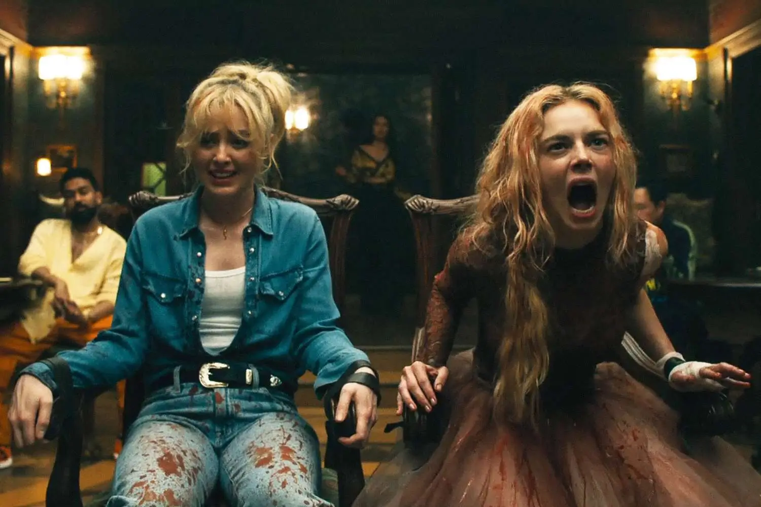 Two women sit restrained in chairs inside a dark, upscale room, their clothes stained with blood as one screams and the other looks distressed, while several figures watch from the background, creating a tense and chaotic horror scene
