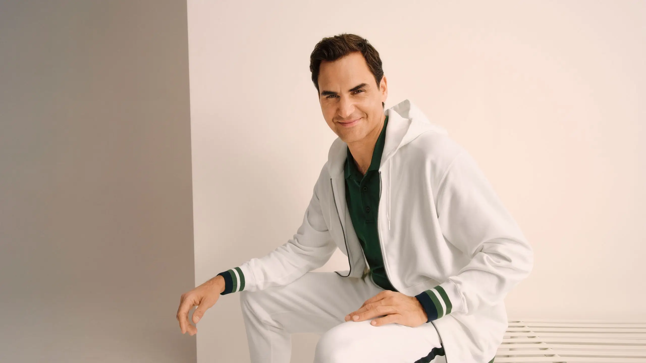 Lifestyle portrait of a male model seated indoors wearing a white UNIQLO tracksuit layered over a green polo shirt, featuring striped cuff details, against a soft neutral backdrop, conveying relaxed elegance and refined sportswear style