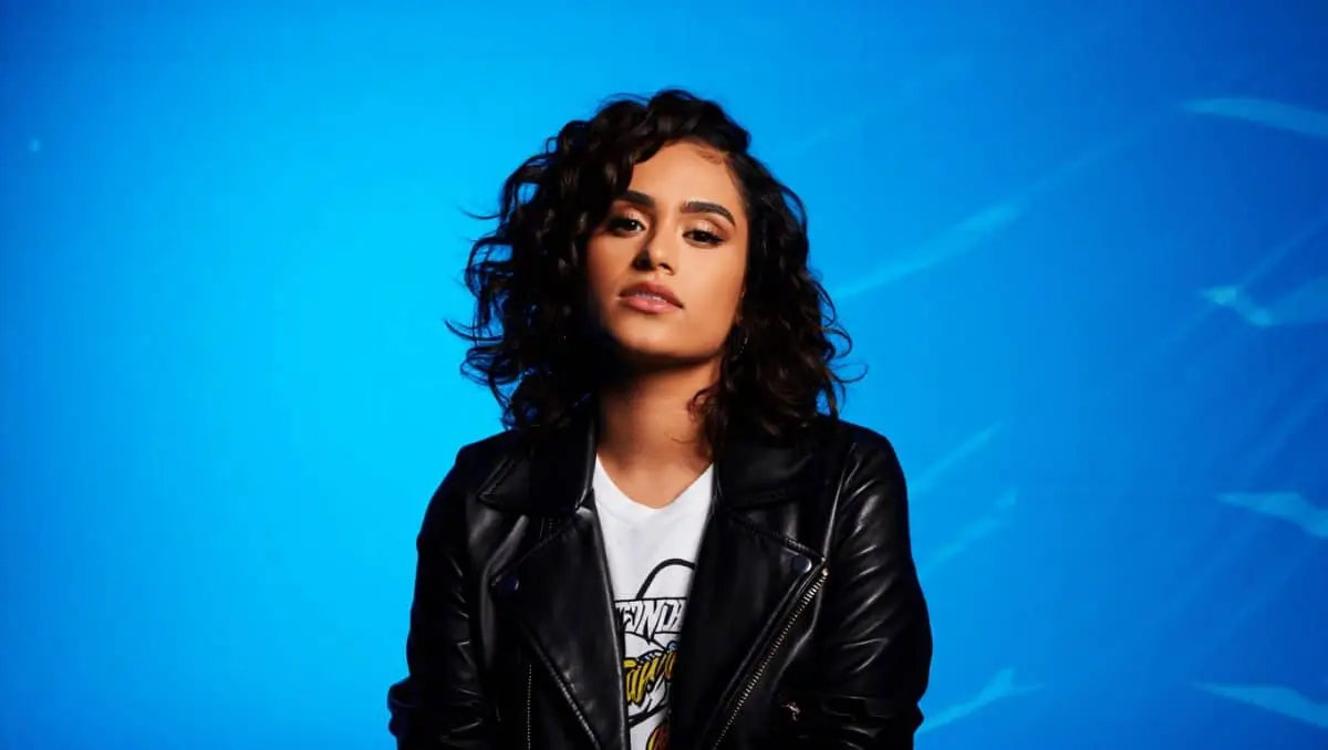 Kehlani in a studio portrait wearing a black leather jacket and white graphic tee, gazing forward with a calm, confident expression against a luminous blue gradient background