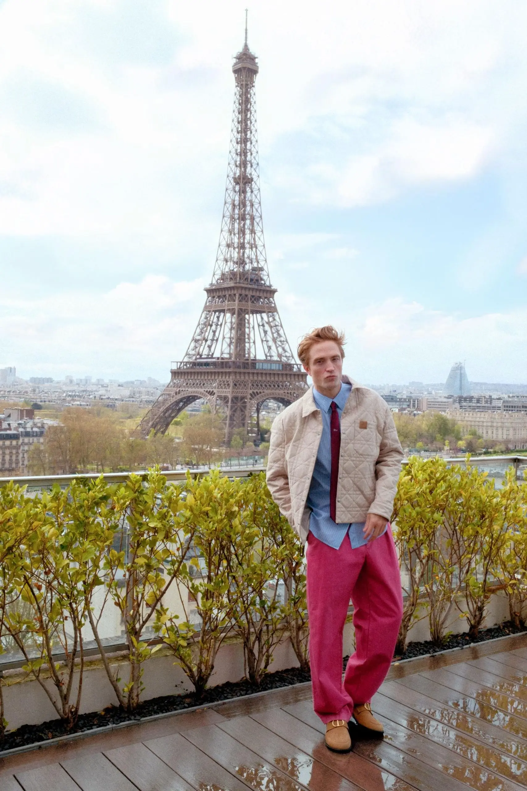 “Robert Pattinson stands on a rain-slick Paris terrace before the Eiffel Tower, wearing a beige quilted Dior jacket over a blue striped shirt with a red tie, paired with loose pink trousers and brown loafers, framed by low greenery and a soft, overcast sky.”