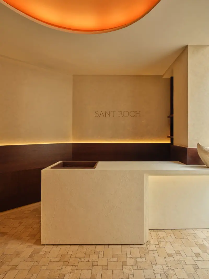 Minimalist reception area at SANT ROCH wellness space in Paris, featuring a sculptural stone desk, warm ambient lighting, and the SANT ROCH logo on a textured plaster wall beneath a glowing circular ceiling light