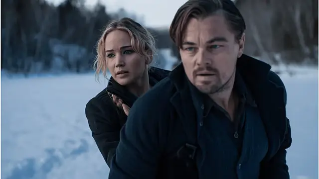 Jennifer Lawrence and Leonardo DiCaprio in winter coats stand close together in a snowy landscape, both looking tense and alert as they glance off-frame, suggesting urgency or danger