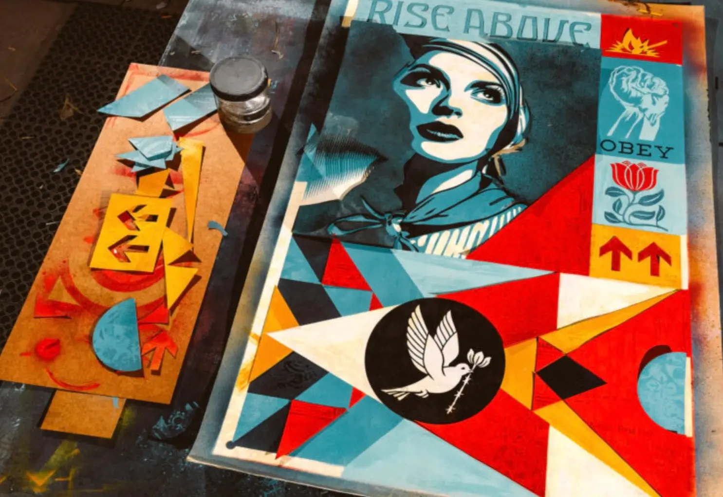Colorful Shepard Fairey stencil artwork on a studio table featuring a stylized female portrait beneath the phrase “Rise Above,” surrounded by geometric shapes, peace symbols, OBEY iconography, and stencil cutouts used in the printmaking process