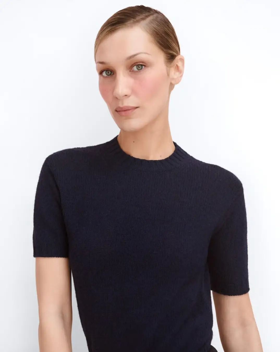 Bella Hadid wearing subtle natural makeup and a navy knit top, posing for Prada Beauty as the brand’s first global ambassador